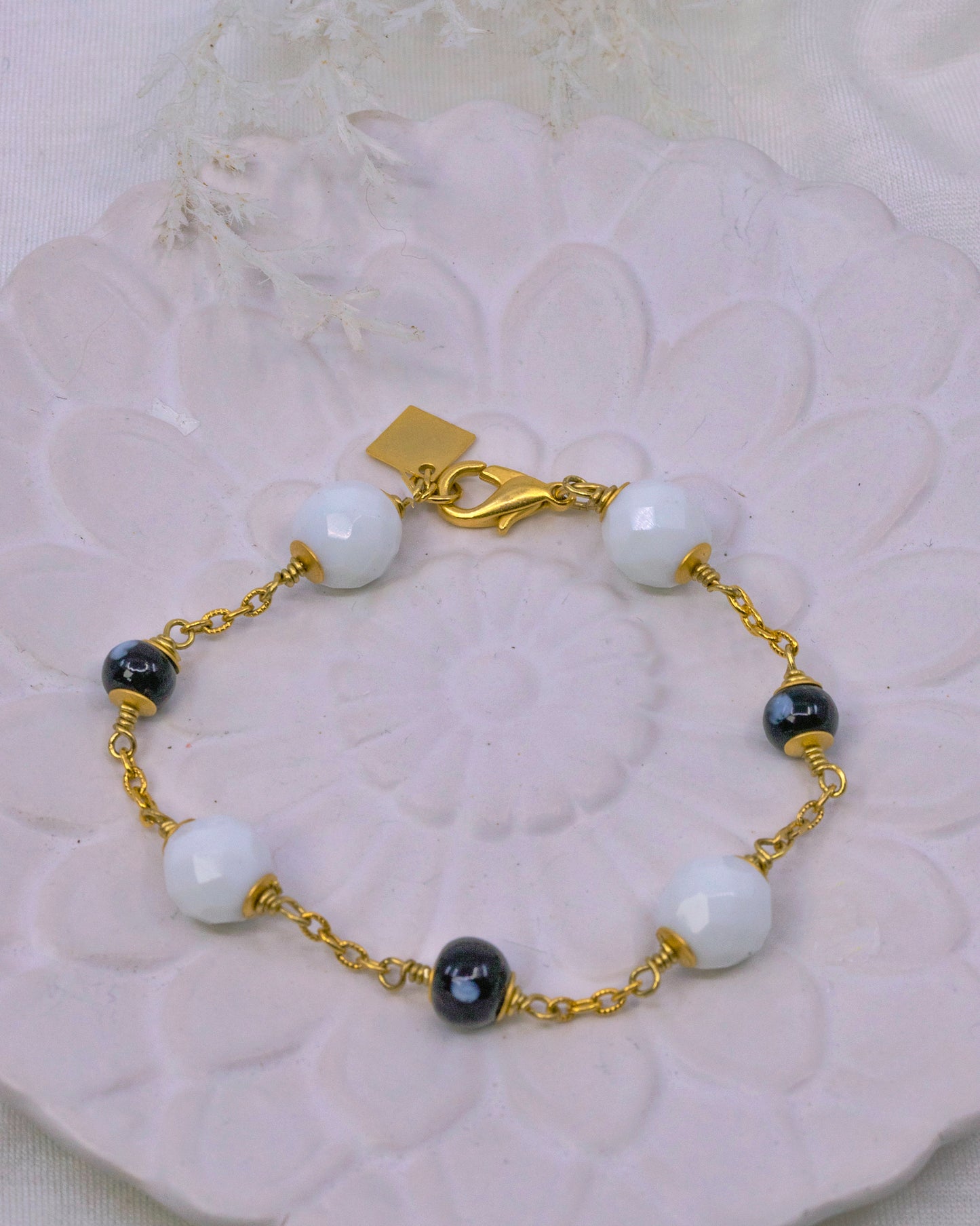 Bracelet with gold chain, white beads, and black stones on a decorative white surface