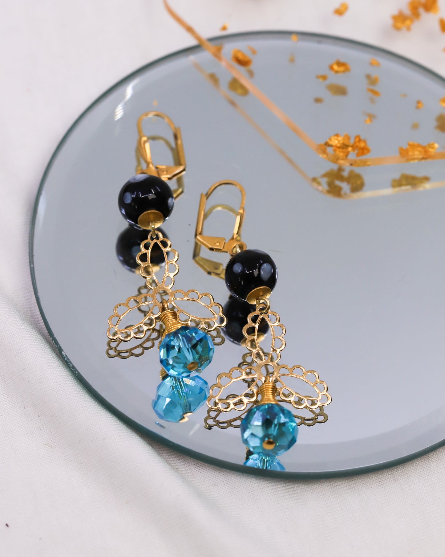 Pair of earrings with gold, black, and blue beads on a reflective surface.