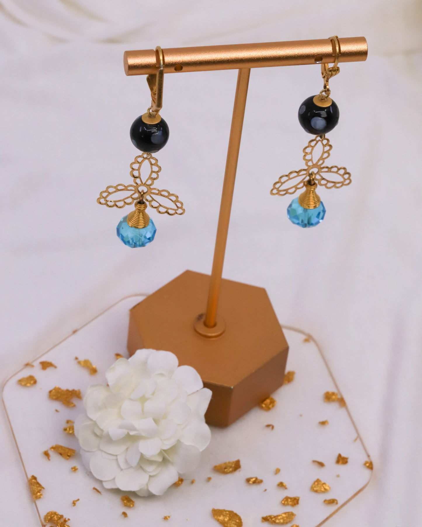 Pair of earrings with black and blue beads on a gold stand with a white flower and gold leaves in the background.