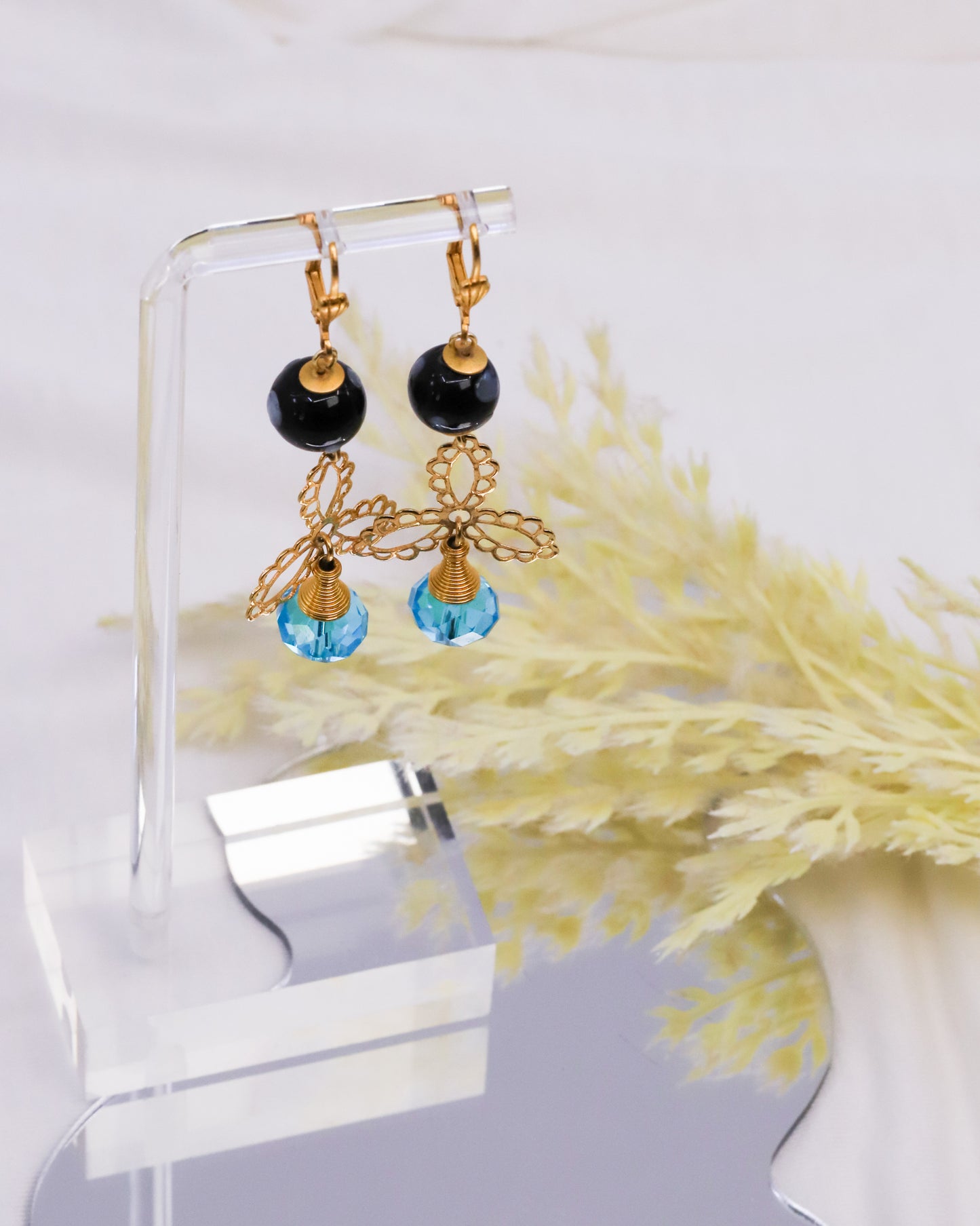 Gold earrings with black and blue beads on a clear stand with dried plants in the background