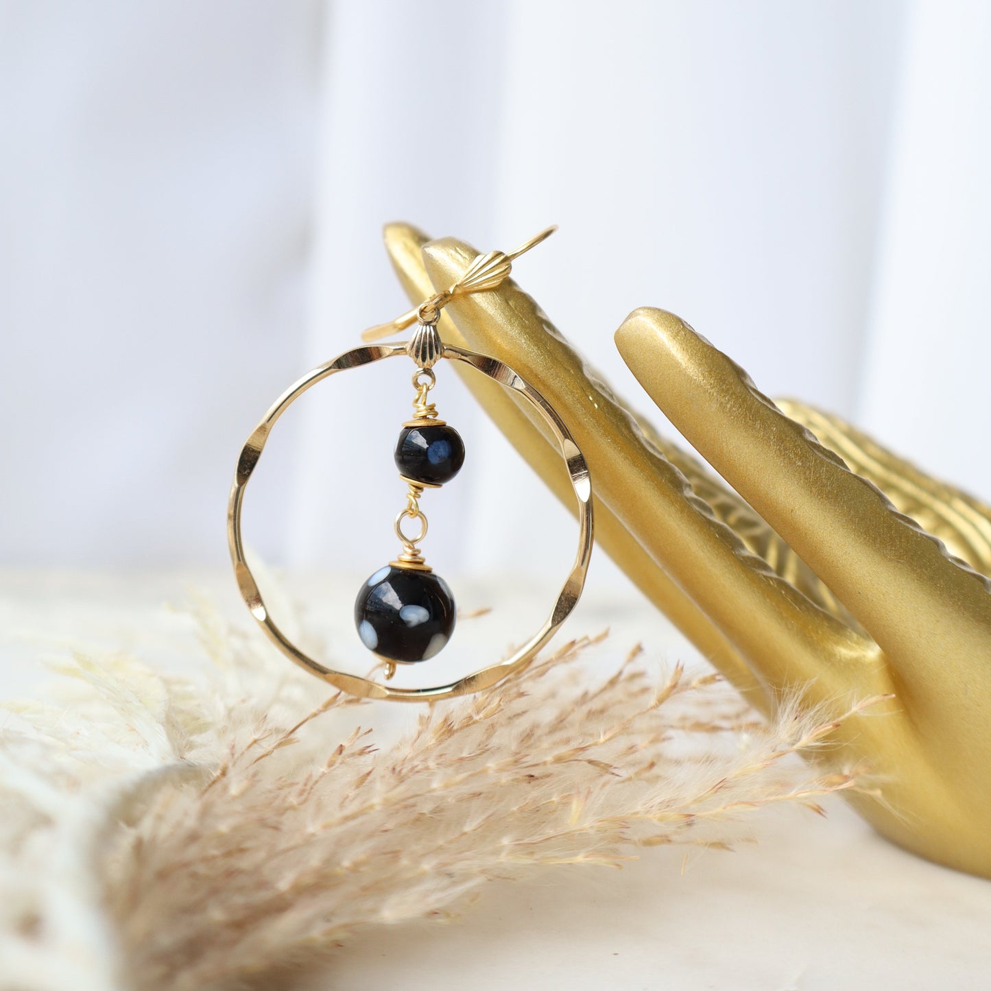 Gold hoop earrings with black beads held by a gold hand against a white background