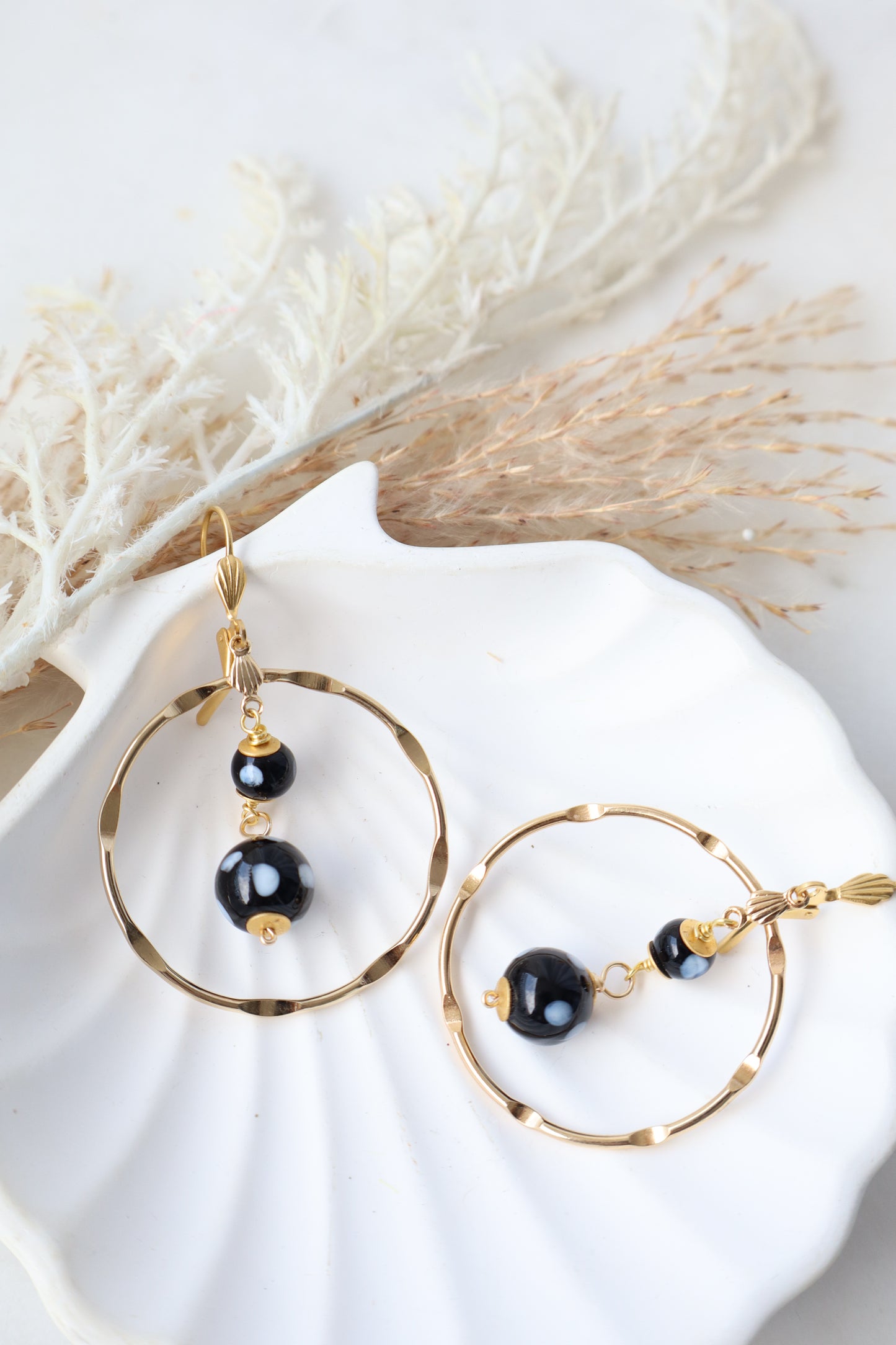 Gold and black earrings on a white shell with dried grasses.