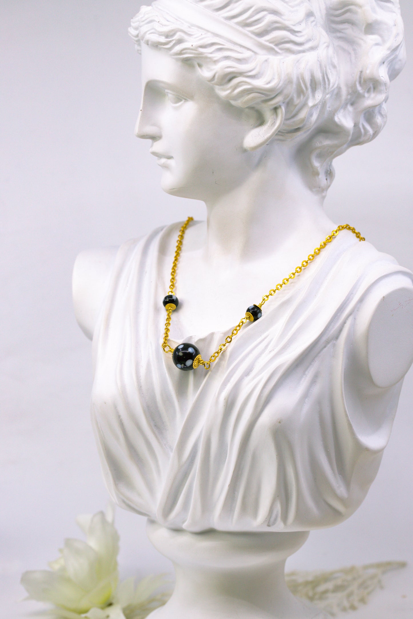 Bust of a classical figure wearing a gold necklace with black beads on a light gray background