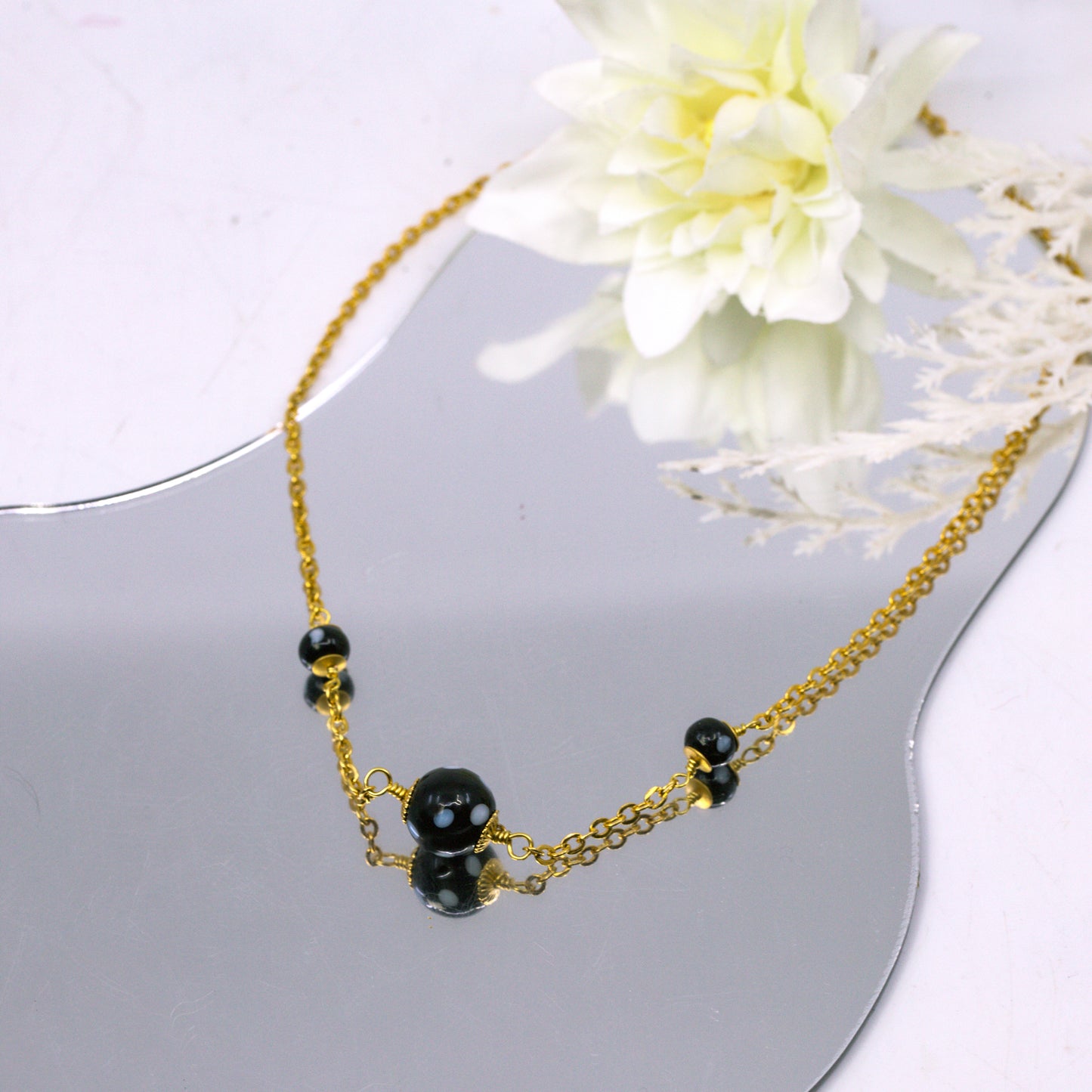 Gold necklace with black beads on a white surface with a white flower in the background