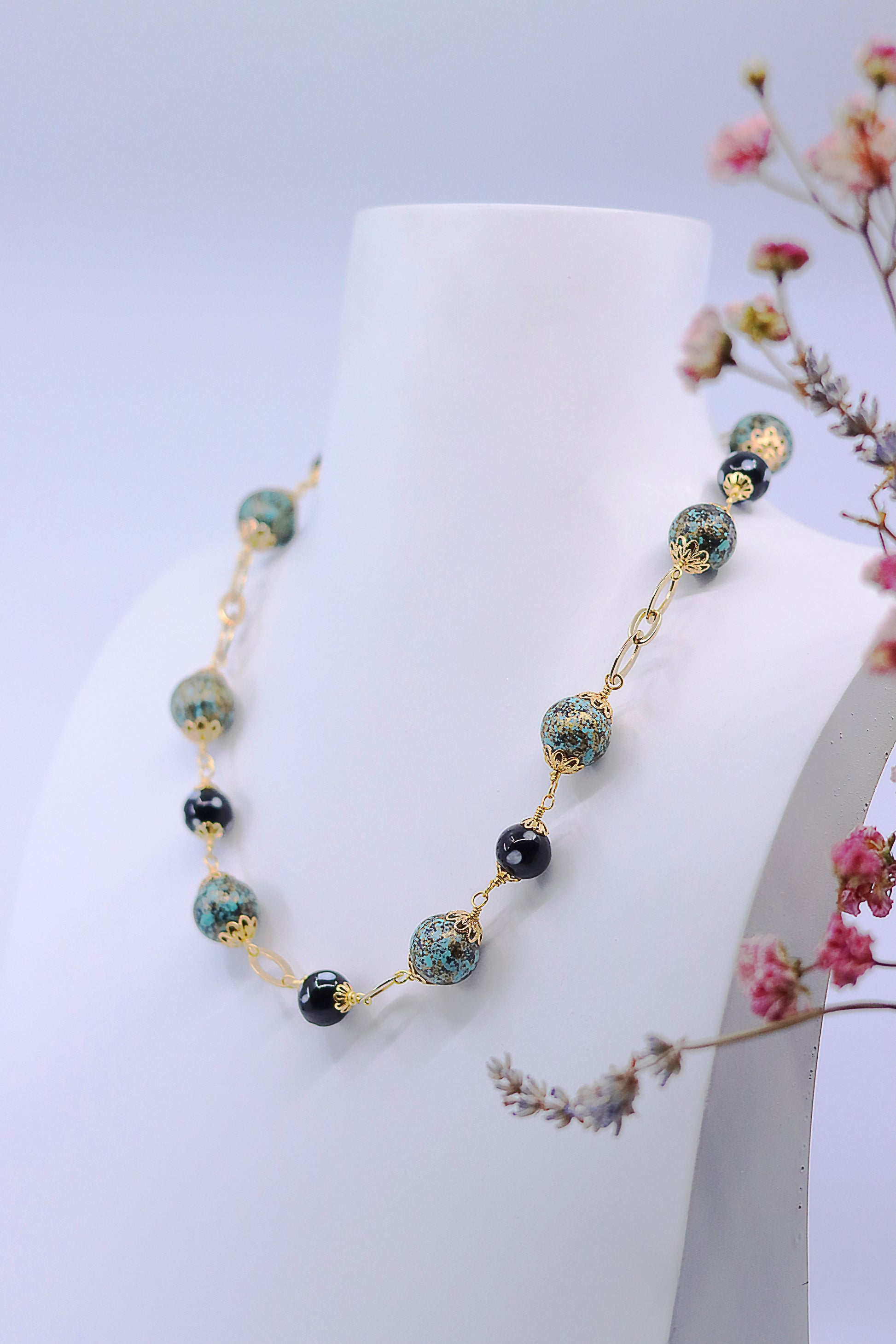 Styled photo of Alcidia Necklace on white bust with floral background, showing bead pattern and gold links