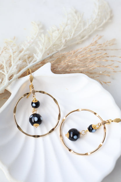 Gold and black earrings on a white shell with dried grasses.
