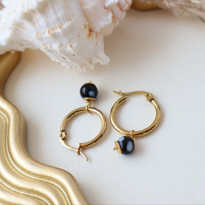 Gold hoop earrings with black stones on a decorative surface with a shell and gold frame.