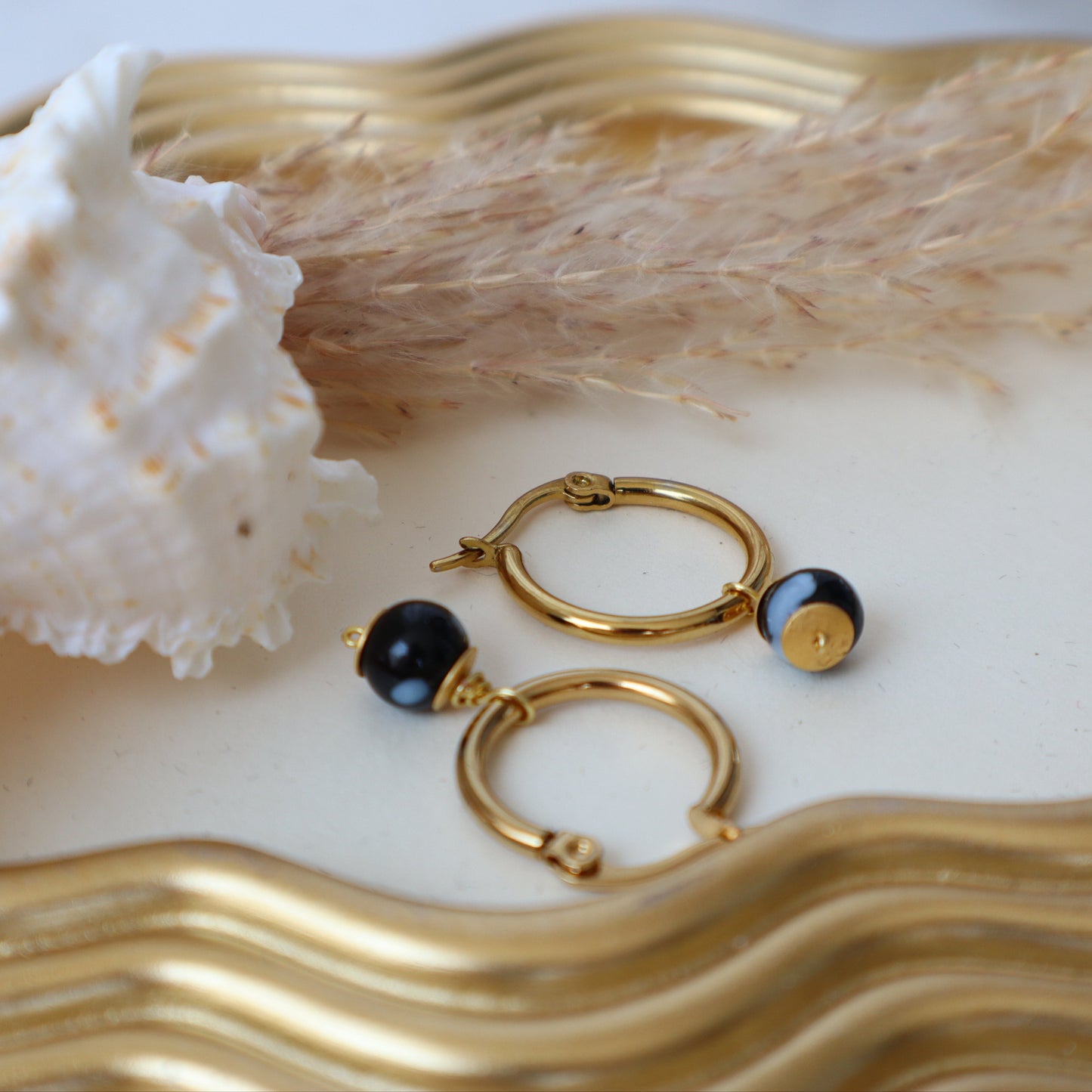 Gold hoop earrings with black stones on a decorative plate with a shell and dried plant.