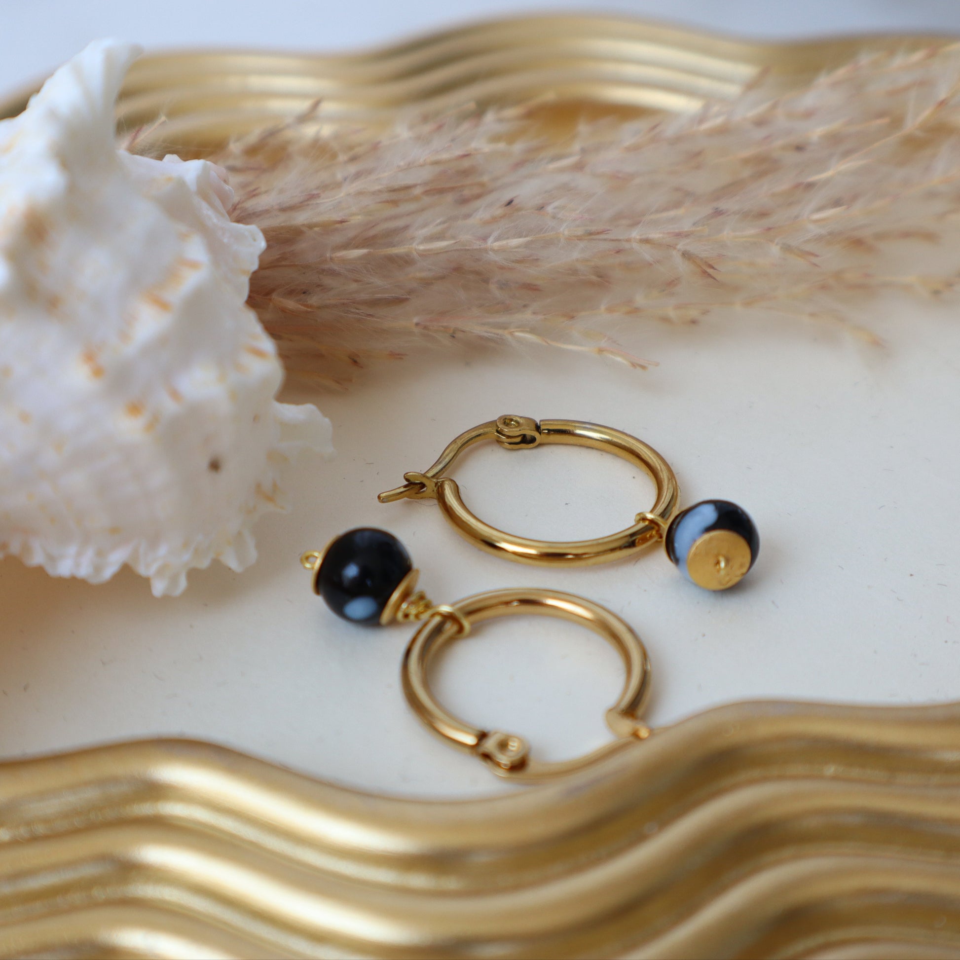 Gold hoop earrings with black stones on a decorative plate with a shell and dried plant.