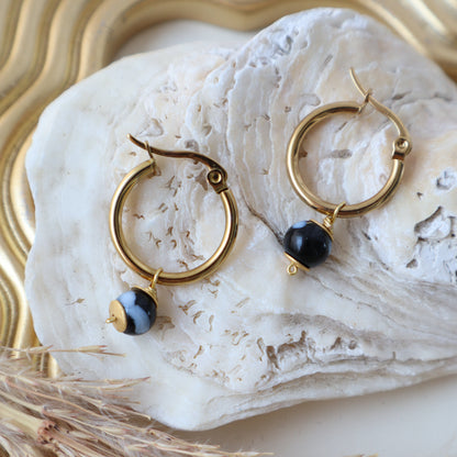 Gold hoop earrings with black stones on a white stone surface with gold and white decorative elements.