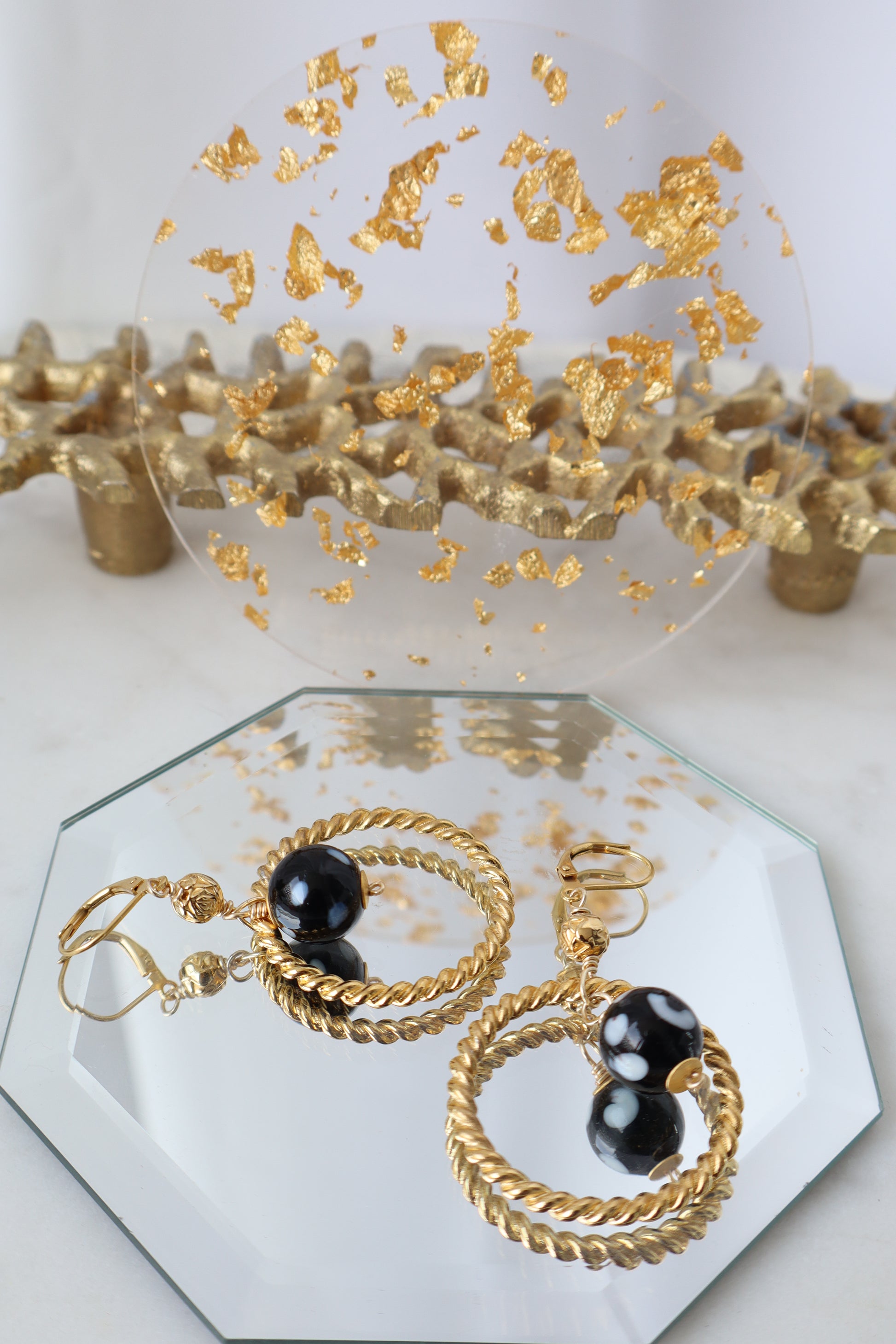 Gold hoop earrings with black stones on a hexagonal mirror with gold leaf accents.