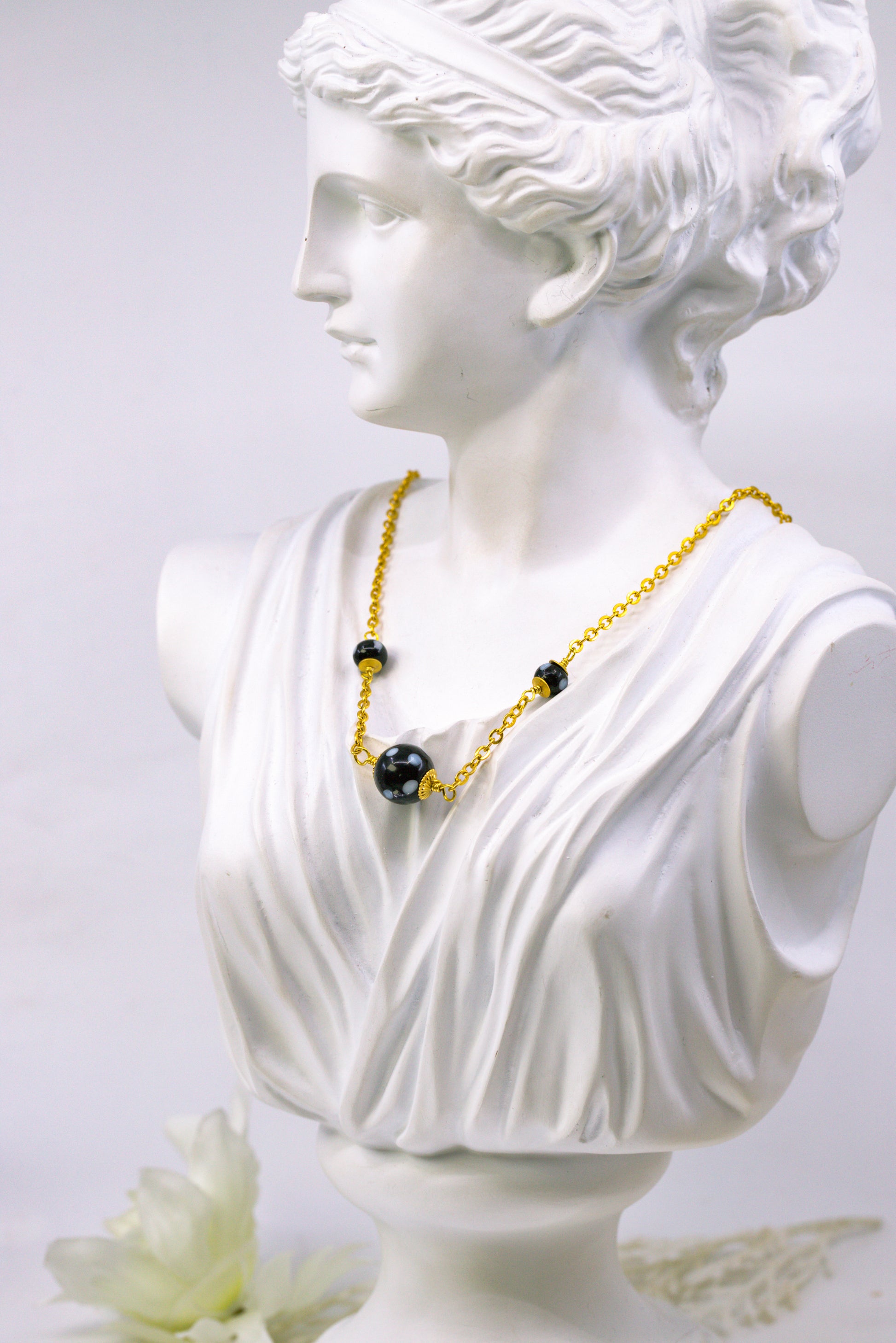 Bust of a classical figure wearing a gold necklace with black beads on a light gray background
