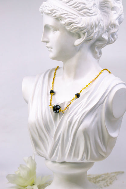 Bust of a classical figure wearing a gold necklace with black beads on a light gray background